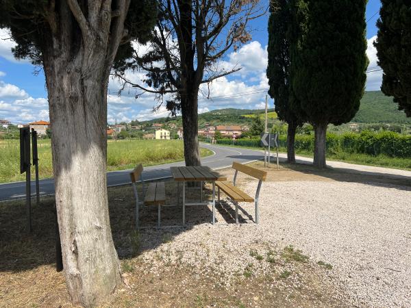 Area di sosta ombreggiata lungo la ciclovia, con tavolo e panchine in legno, circondata da cipressi.